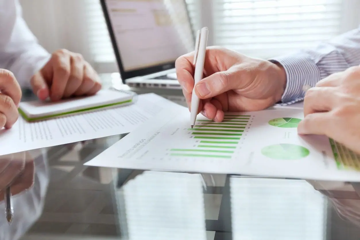 Two professionals analyzing financial documents with graphs and charts on a desk in a bright office.