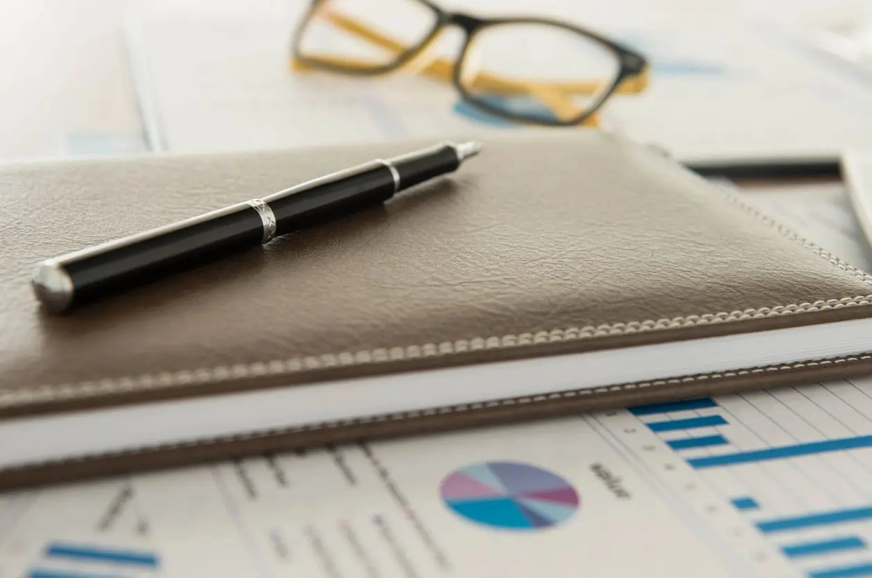 A close-up view of a desk with a brown leather notebook, black pen, eyeglasses, and documents with charts.