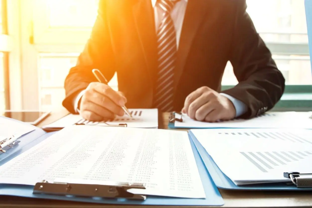 A businessman in a suit reviews documents at a desk with clipboards and reports, sunlight streaming in from the window.